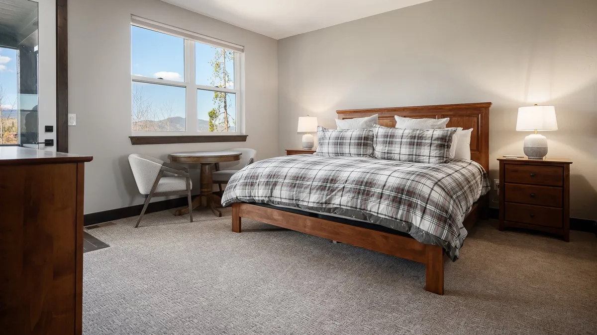 A spacious bedroom featuring a large bed with plaid bedding, a wooden side table, and a window revealing serene mountain views. The decor emphasizes warmth and tranquility, with premium bedding and cozy touches apparent.