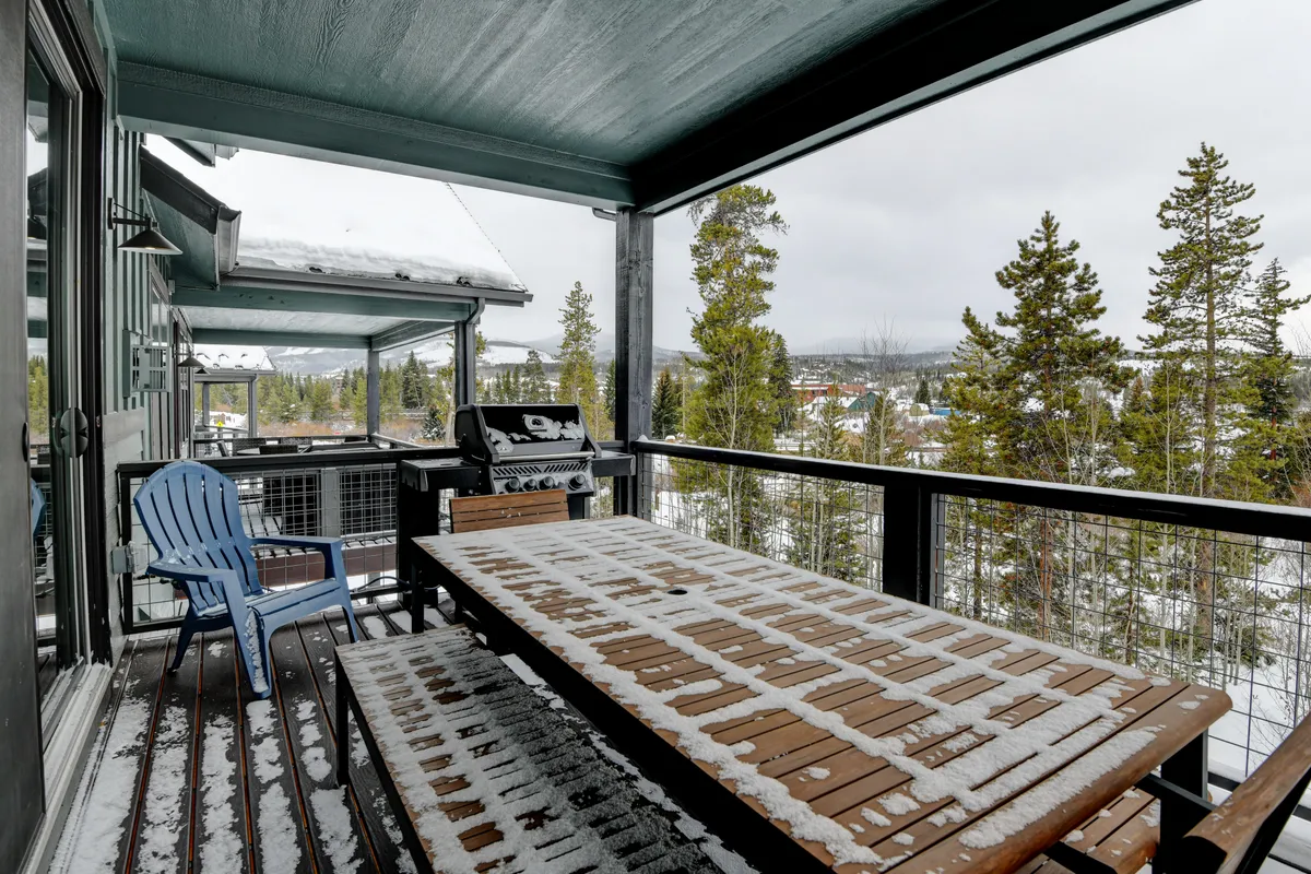 A welcoming exterior deck features a wooden dining table set for a meal, blue Adirondack chairs, a grill, and a snowy woodland backdrop. The inviting space is ideal for sharing food, stargazing, or relaxing with loved ones in the fresh mountain air.
