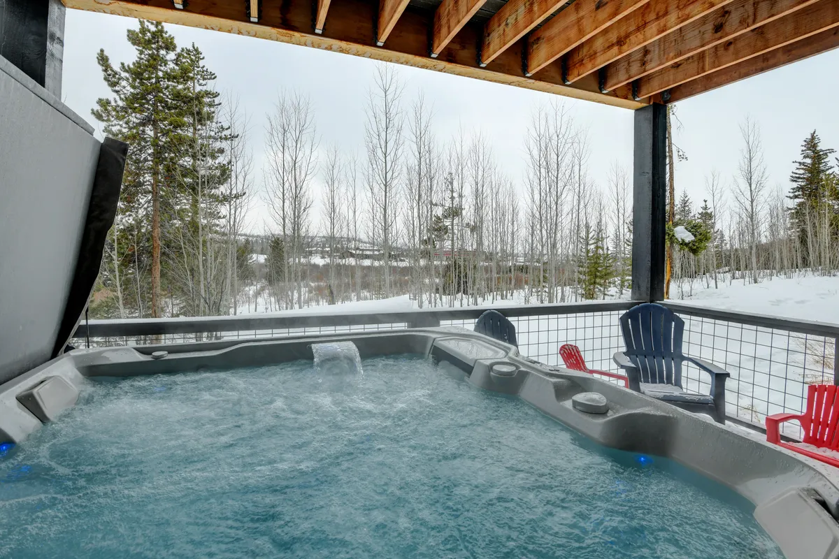 This image depicts a snow-covered lower patio featuring a private hot tub surrounded by tall trees. Adirondack chairs are arranged around the hot tub, inviting relaxation while snow gently falls, creating a peaceful winter retreat. The setting exudes tranquility and connection with nature, perfect for unwinding after outdoor adventures.