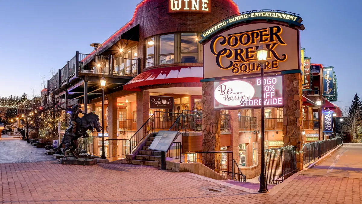 The image showcases the vibrant Cooper Creek Square, with its recognizable sign, outdoor seating areas, staircase, and a horse sculpture. The atmosphere is lively and festive, hinting at a bustling downtown experience with dining, shopping, and socializing.
