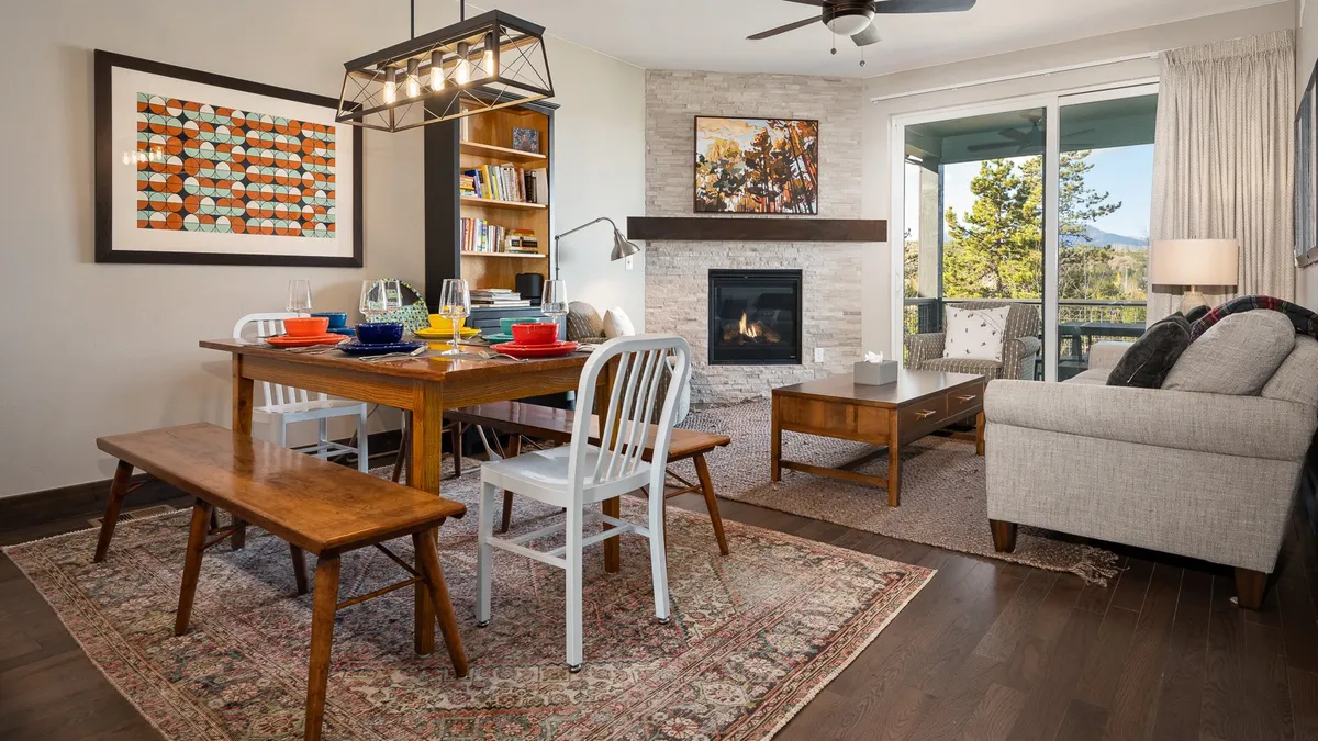 An open-plan interior with a dining area, fireplace, sitting area, and a bookshelf. The warm lighting and comfortable furnishings create a space ideal for game nights, reading, or simply relaxing by the fire.