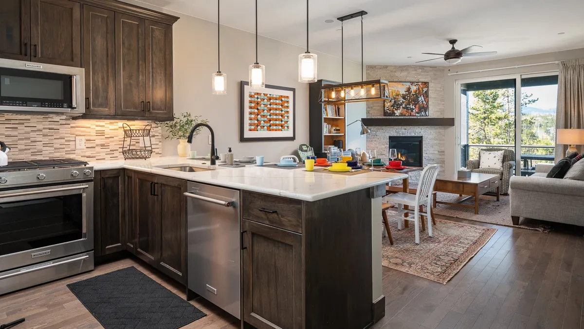 An open-concept interior featuring a kitchen island and dining table, with a cozy fireplace and large windows in the background. The space exudes warmth and comfort, inviting group meals, celebrations, and morning rituals in a setting that blends kitchen and living areas seamlessly.