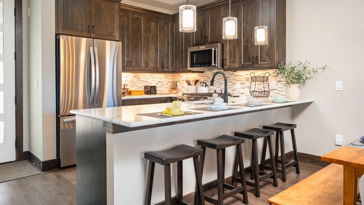 A modern kitchen island with comfortable bar stools sits beneath pendant lighting, flanked by stainless steel appliances and a vibrant tile backsplash. The inviting breakfast bar faces into open living spaces, creating a seamless flow for socializing and shared meals.