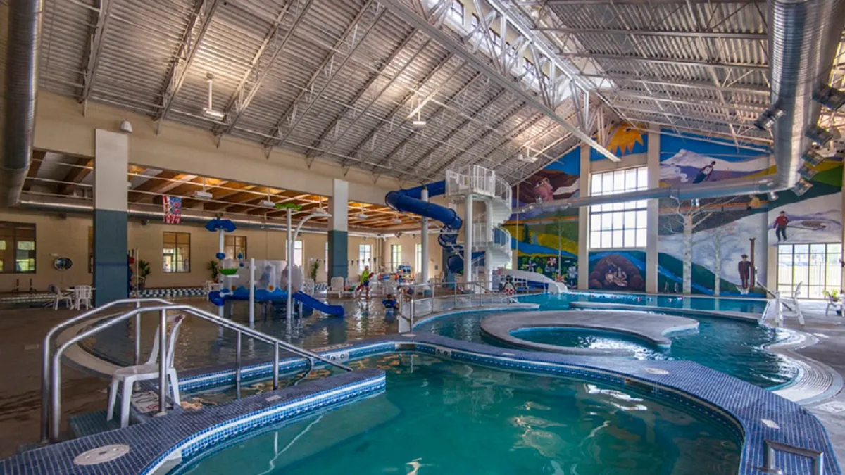 Inside a lively recreation center, children splash and laugh in a large swimming pool while others zip down a colorful waterslide. Bright murals add to the energetic, family-friendly atmosphere, capturing the excitement of group play and aquatic adventures.