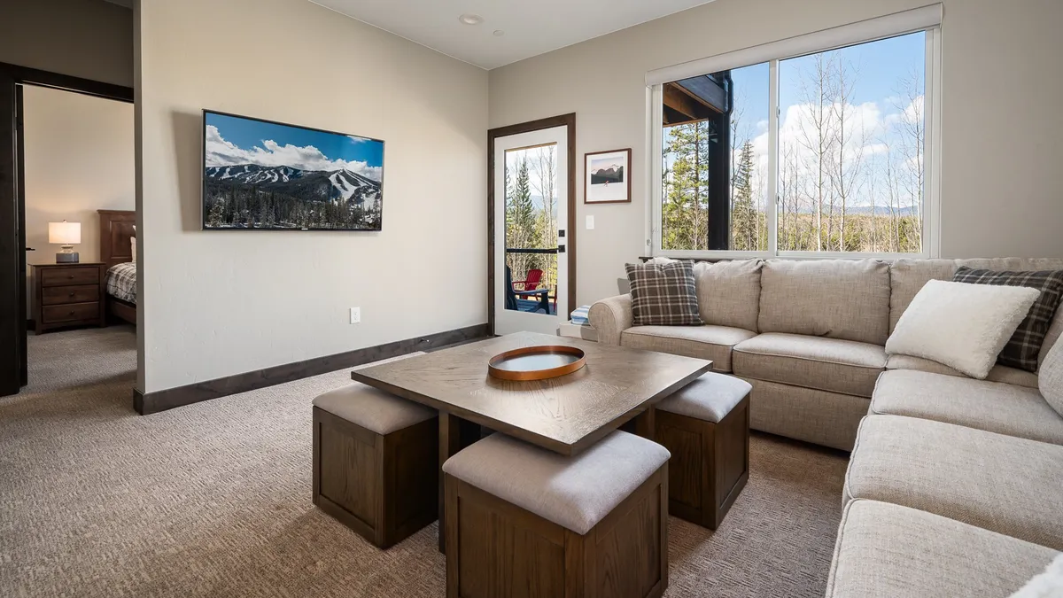 A spacious, inviting living area featuring a large wall-mounted HD Smart TV, a comfortable sectional sofa, a coffee table with a decorative bowl, and expansive windows offering breathtaking mountain views. The space is filled with natural light, creating a warm and welcoming atmosphere ideal for gathering and relaxing.