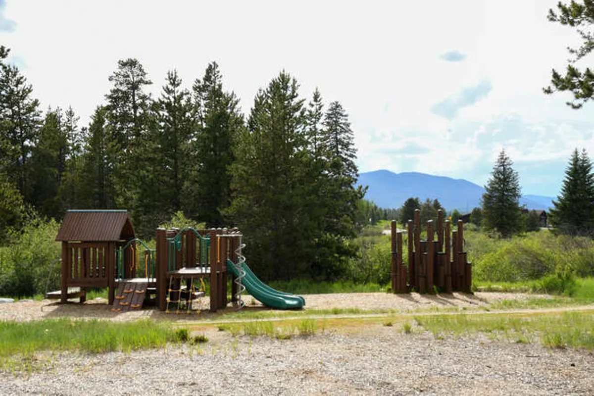 A vibrant playground structure with slides sits nestled among tall trees and a stunning mountain backdrop. The area is buzzing with cheerful children and families enjoying outdoor activities, evoking a sense of adventure and togetherness in a natural setting.