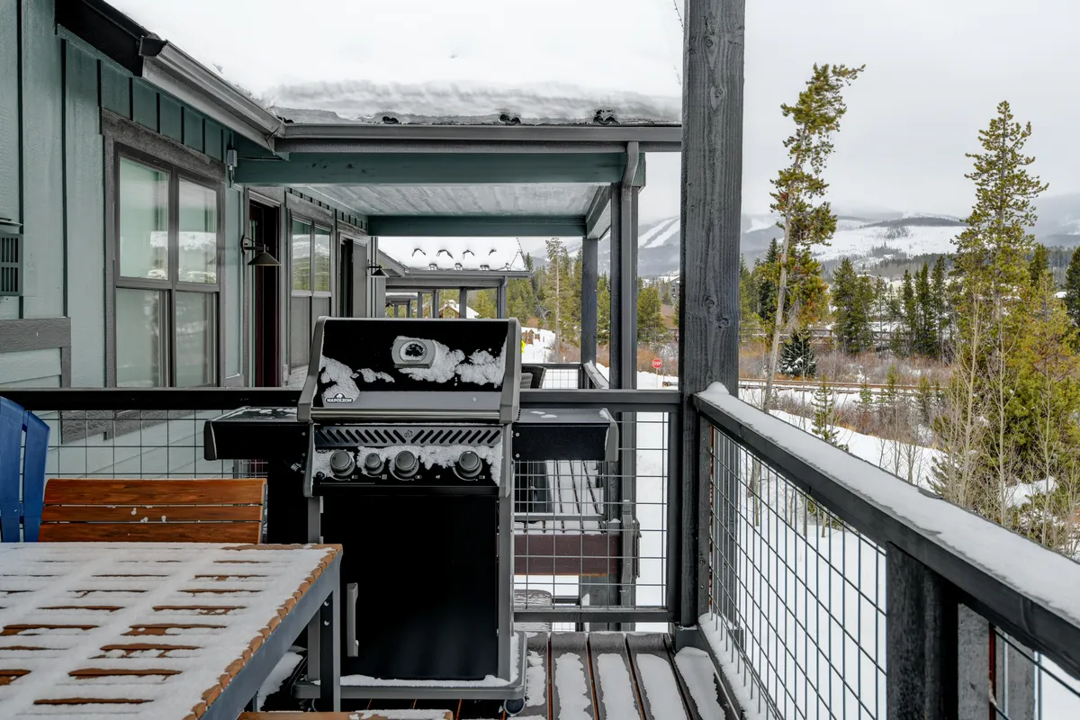 A cozy winter deck scene with a wooden table, grill, and snow-covered surfaces, set against a sweeping mountain view. The ambiance is warm and inviting—perfect for après-ski gatherings, outdoor dining, or enjoying hot drinks as the sun sets over the snowy landscape.