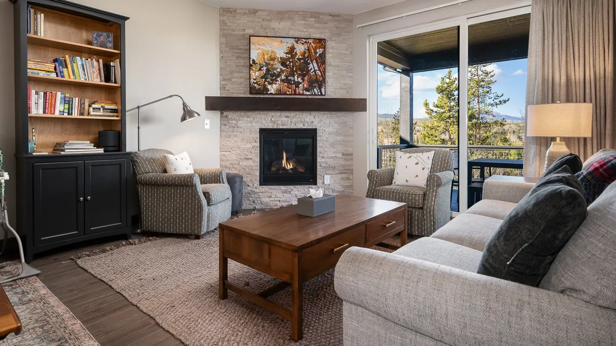 A warm, inviting living room with a crackling fireplace, built-in bookshelves, stylish coffee table, and expansive windows framing mountain views. The space evokes coziness, perfect for unwinding, reading, or gathering with friends and family.